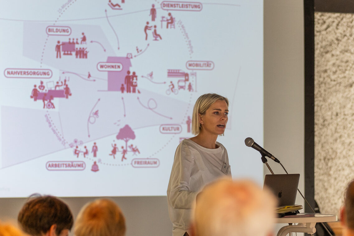 Eine Frau steht vor einer Gruppe von Frauen und Männern an einem Pult und spricht. Hinter ihr wird ein Bild ihres Konzeptes gezeigt.