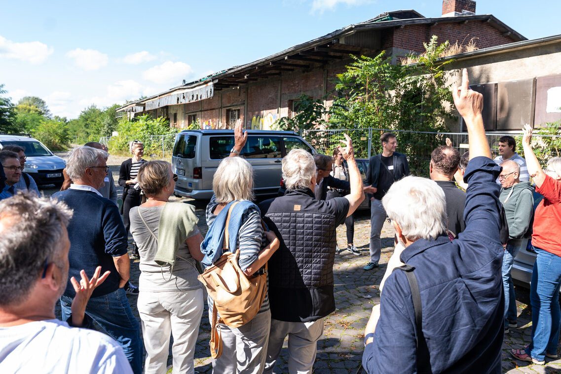 Eine Gruppe von Frauen und Männern steht vor einer verfallenen Güterhalle. Einige Personen heben die Hand.
