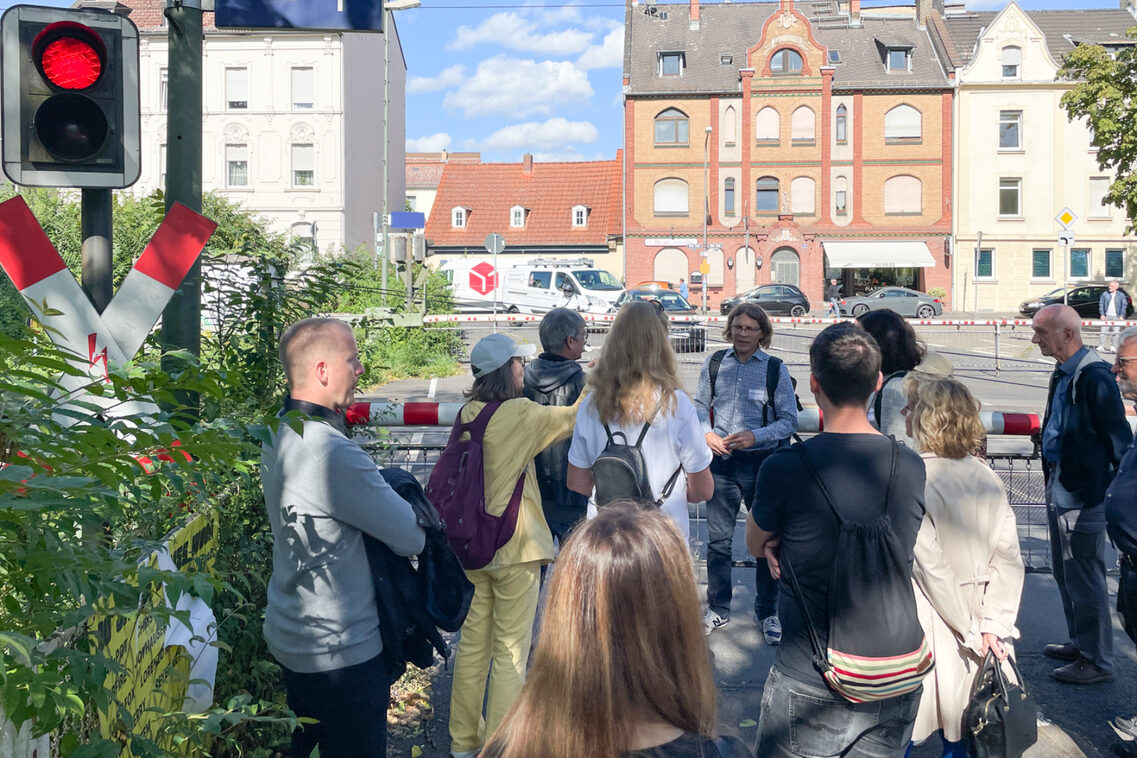 Eine Gruppe von Frauen und Männern steht an einem geschlossenen Bahnübergang und diskutiert.
