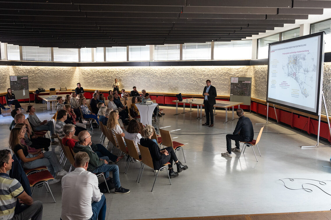 Bürgerinnen und Bürger sitzen in einem großen Saal und hören der Präsentation eines Mannes zu.