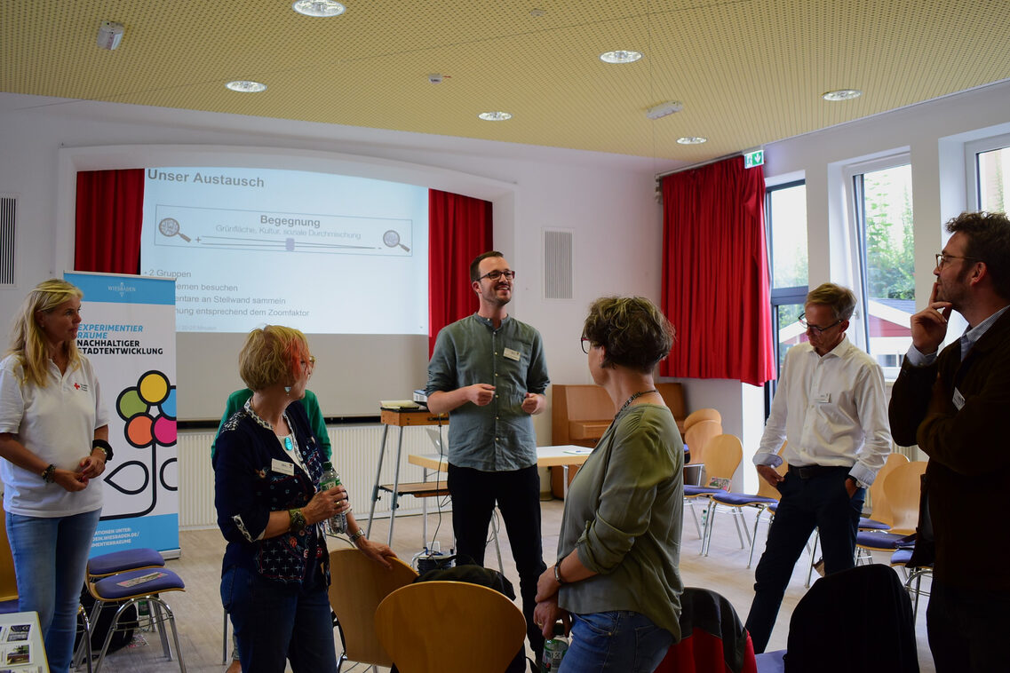 Eine Gruppe von Frauen und Männern steht in einem Saal mit Stühlen und diskutiert. Im Hintergrund ist eine Präsentation zu sehen.