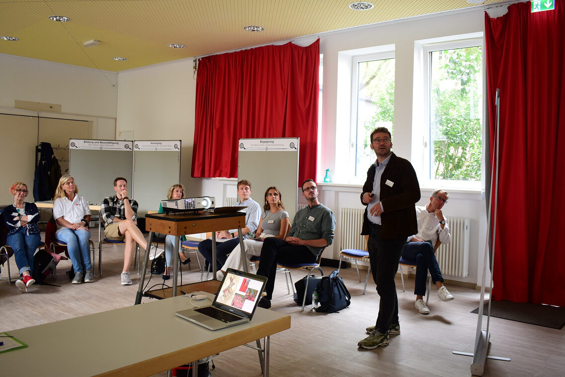 Eine Gruppe von Frauen und Männern schaut auf eine Präsentation. Die Gruppe befindet sich in einem Saal mit Tischen sowie Stellwänden und sitzt in einem Halbkreis auf Stühlen. Ein Mann steht und präsentiert.