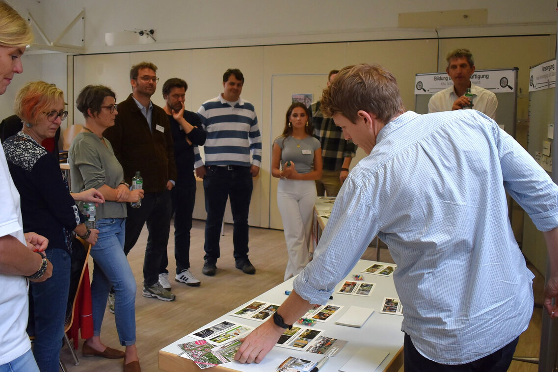Ein Mann zeigt einer Gruppe von Frauen und Männern ausgedruckte Bilder, die vor ihm auf einem Tisch ausliegen.