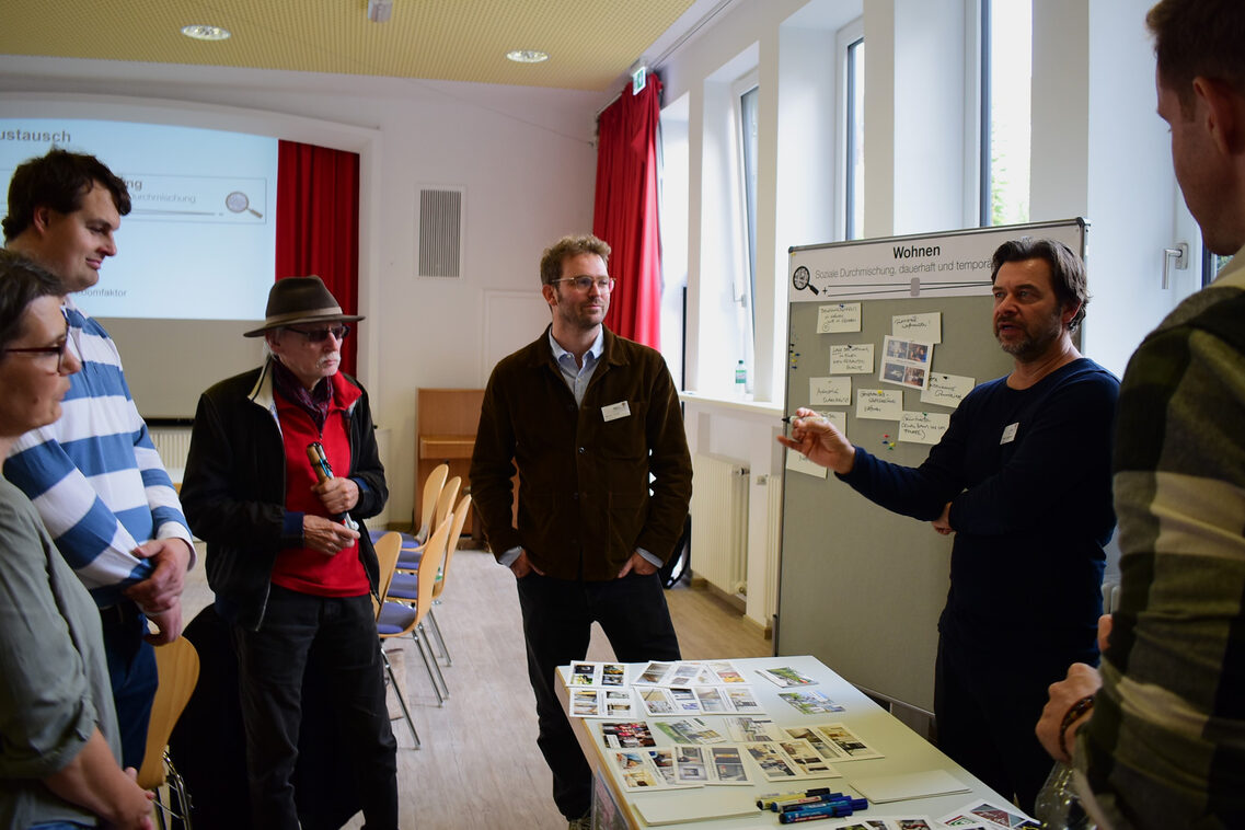 Fünf Männer und eine Frau stehen vor einer Stellwand mit Zetteln und Bildern. Zwischen ihnen steht ein Tisch mit gedruckten Bildern.