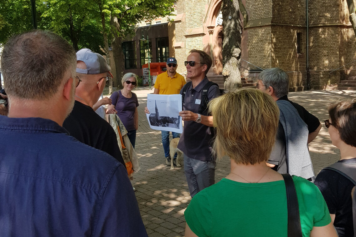 Eine Gruppe von Frauen und Männern steht auf einem Platz vor einer Kirche. Ein Mann mit Sonnenbrille zeigt der Gruppe ein historisches Foto.