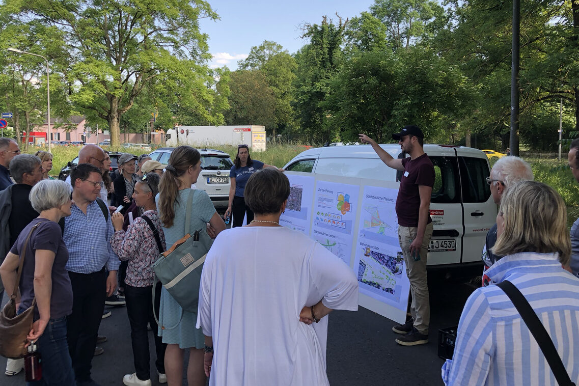 Ein Mann zeigt einer Gruppe von Frauen und Männern Pläne. Die Gruppe befindet sich auf dem Bahnhofsvorplatz vor parkenden Autos.