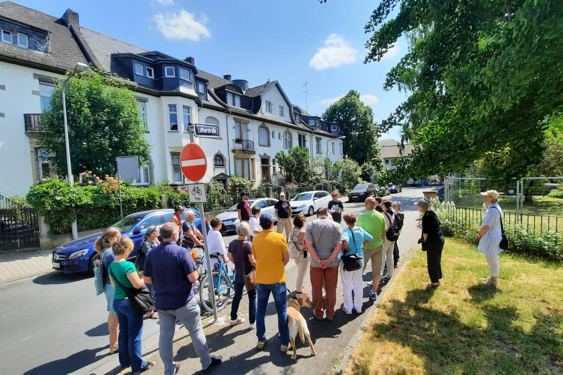 Eine Gruppe von Frauen und Männern steht auf einem Bürgersteig vor Wohnhäusern.
