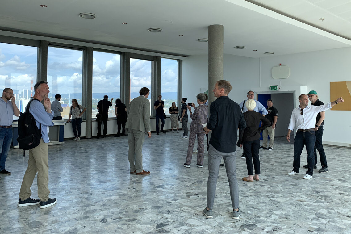 Eine Menschengruppe besichtigen einen großen Raum aus dessen Fenster man einen Blick über Wiesbaden hat.