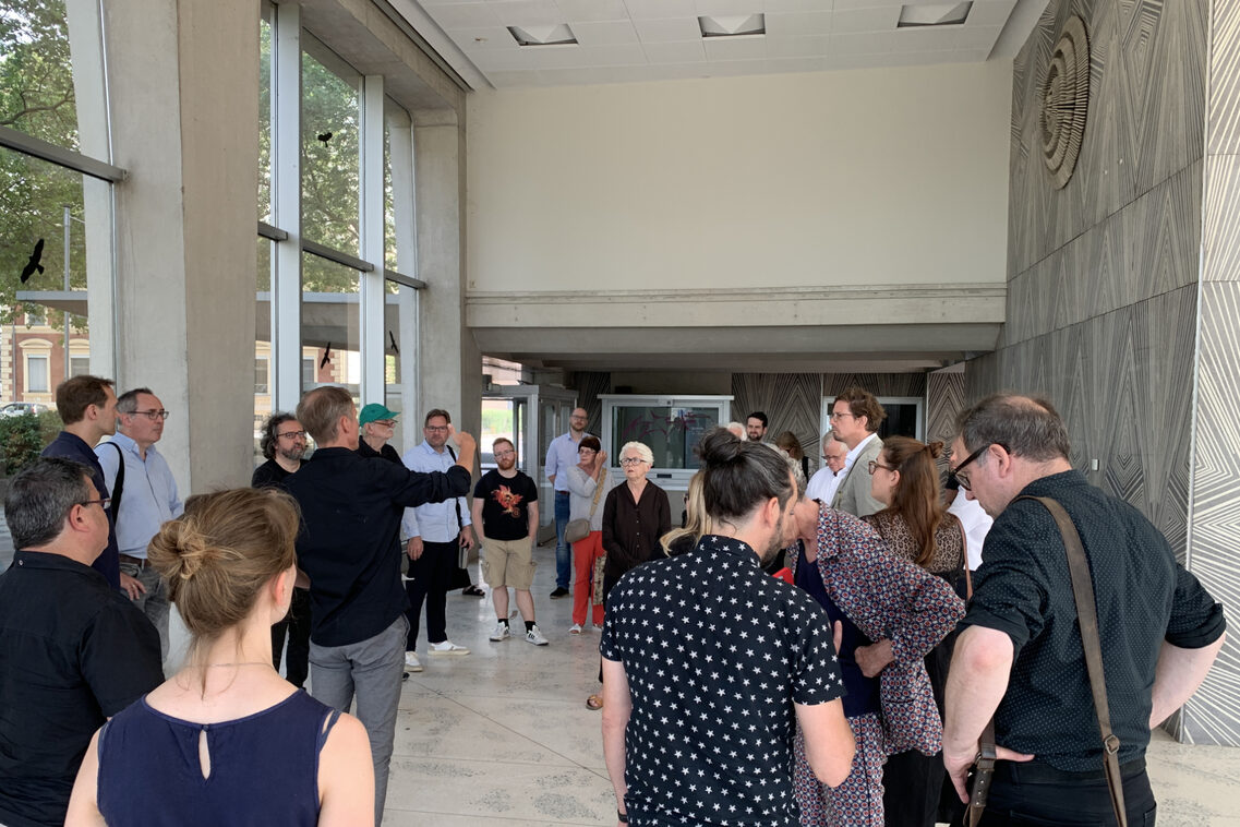 Eine Menschengruppe ist in einem hellen Foyer mit einander im Gespräch.