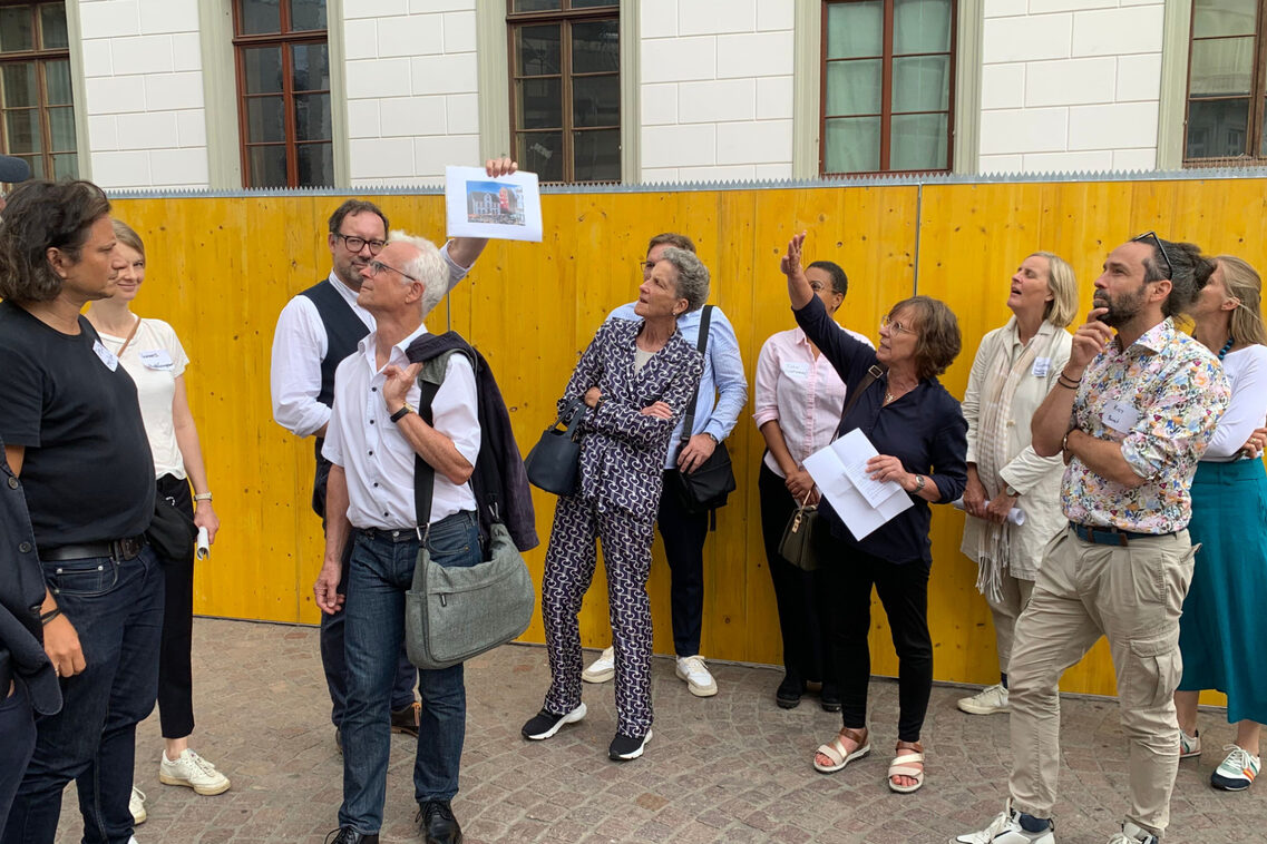 Einer Menschengruppe wird vor einem Bauzaun ein Nachbargebäude präsentiert.