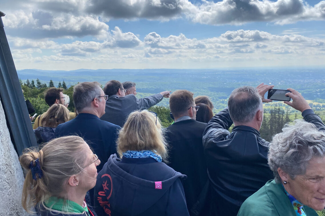 Eine Menschengruppe blickt von dem Umgang eines Turms im Wald auf Wiesbaden.