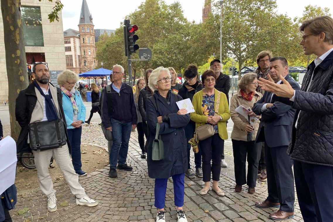Einer Menschengruppe wird an einem Platz ein Bauvorhaben präsentiert.