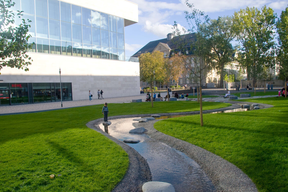 Auf dem Platz der deutschen Einheit fließt ein freigelegter Bach, ein Kind spielt am Wasser.