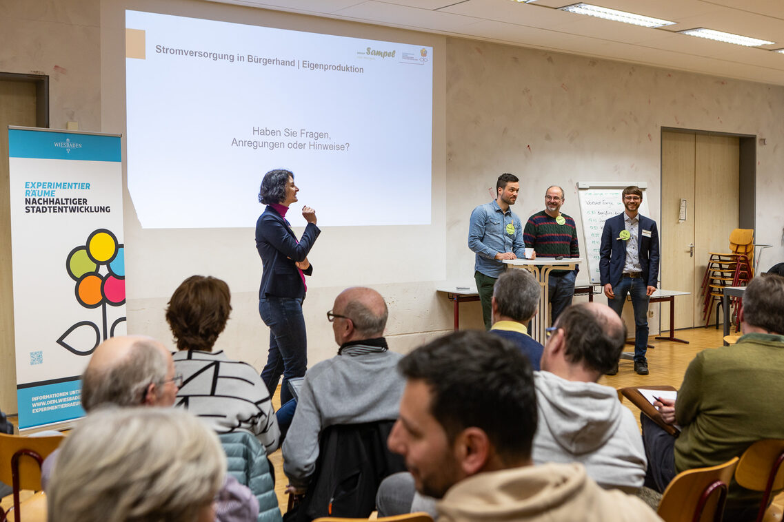 Mehrere Menschen sitzen in einem Raum und hören einem Vortrag zu, der auf einer Leinwand gezeigt wird.