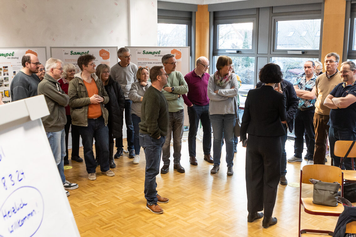 Mehrere Menschen stehen in einem Raum verteilt und werden von einer Frau in der Mitte befragt, im Hintergrund sieht man verschiedene Informationstafeln.