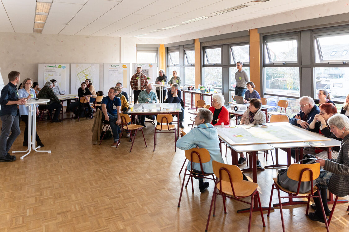 In a large room, several people sit together in table groups and listen to a lecture; plans are laid out on the tables.