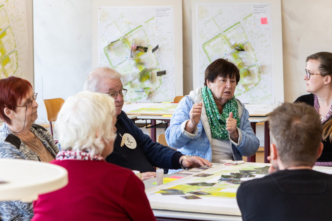 Six people are sitting around a table with plans and colorful cards and an elderly lady is talking.