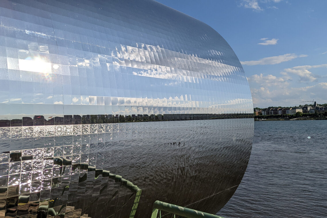 The sculpture's pixel sheets reflect the water and the sky.