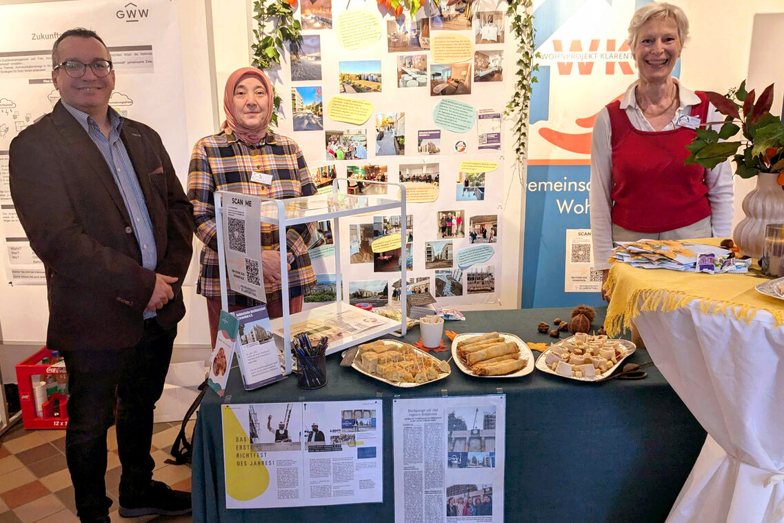 Drei Personen stehen hinter bzw. neben einem dekorierten Tisch mit Infomaterialien und verschiedenen Teiggerichten. Sie lächeln in die Kamera. Im Hintergrund sind eine Pinnwand mit Bildern und ein Infobanner.