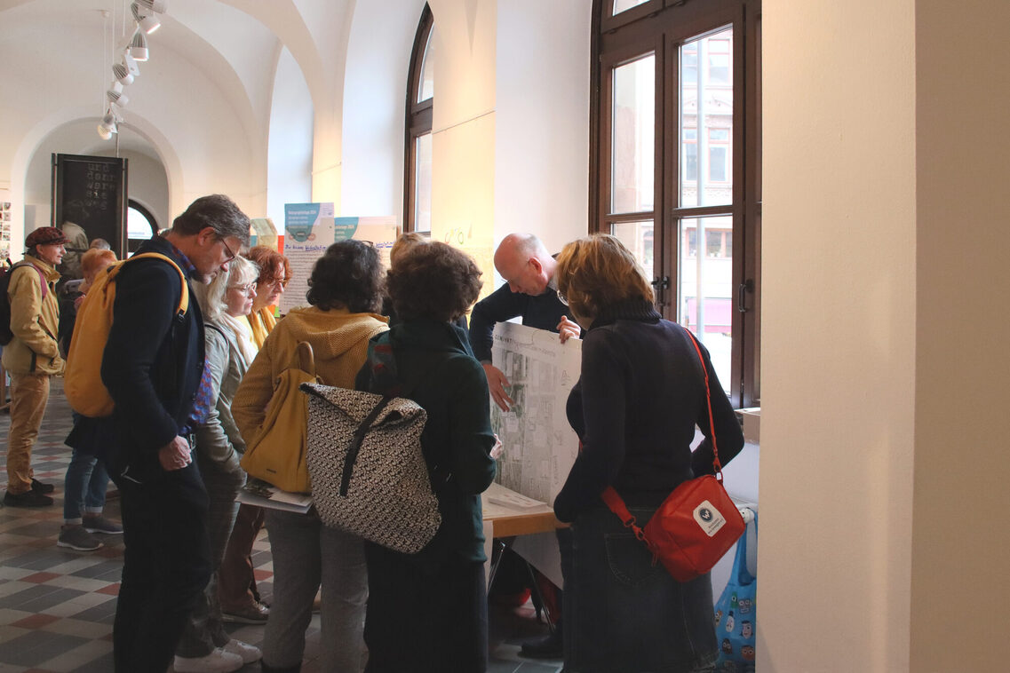 Mehrere Personen informieren sich an einem Tisch und blicken auf einen ca. Din A0 großen Plan, den ein Mann mit Halbglatze in den Händen haltend präsentiert.