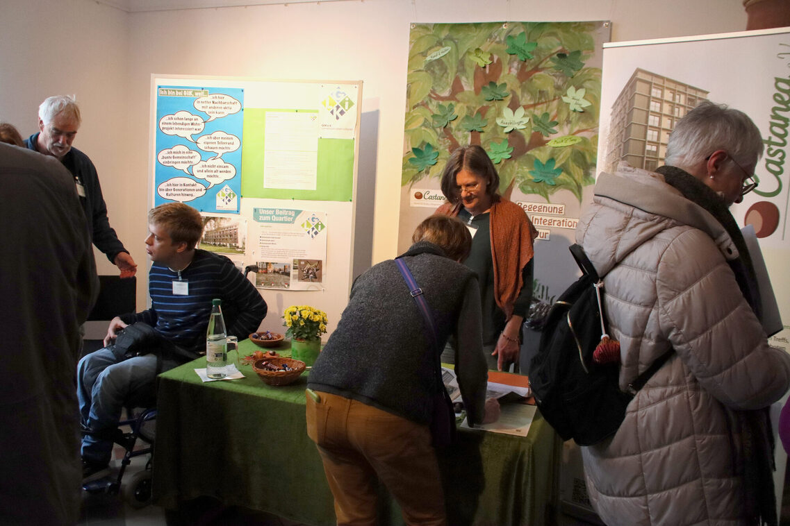 Mehrere Personen stehen um einen Informationsstand herum. Eine Person sitzt im Rollstuhl. Sie bemerken die Kamera nicht. Im hintergrunf sind verschiedene Banner zu sehen, auf einem ist ein Comic abgebildet. Auf einem anderen ist der Schriftzug „Castanea“ zu erkennen.