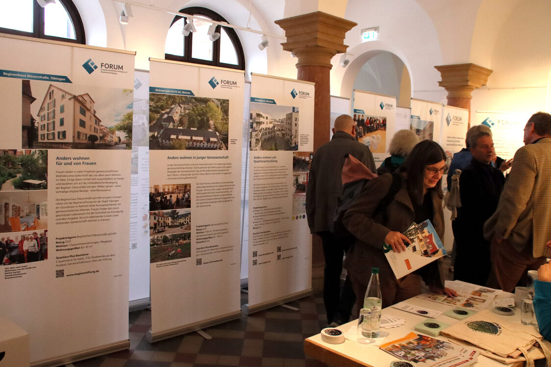 A row of floor-to-ceiling information banners in a room with columns and a checkered stone floor. On the right, several people talking, studying the banners and a person helping themselves at a table with information material.
