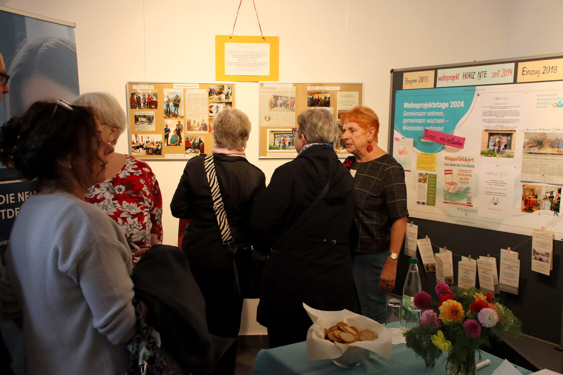 Five people stand facing away from the camera in front of several pinboards and look at the pictures hanging on them. In the foreground is a table with flowers and a basket of bread.