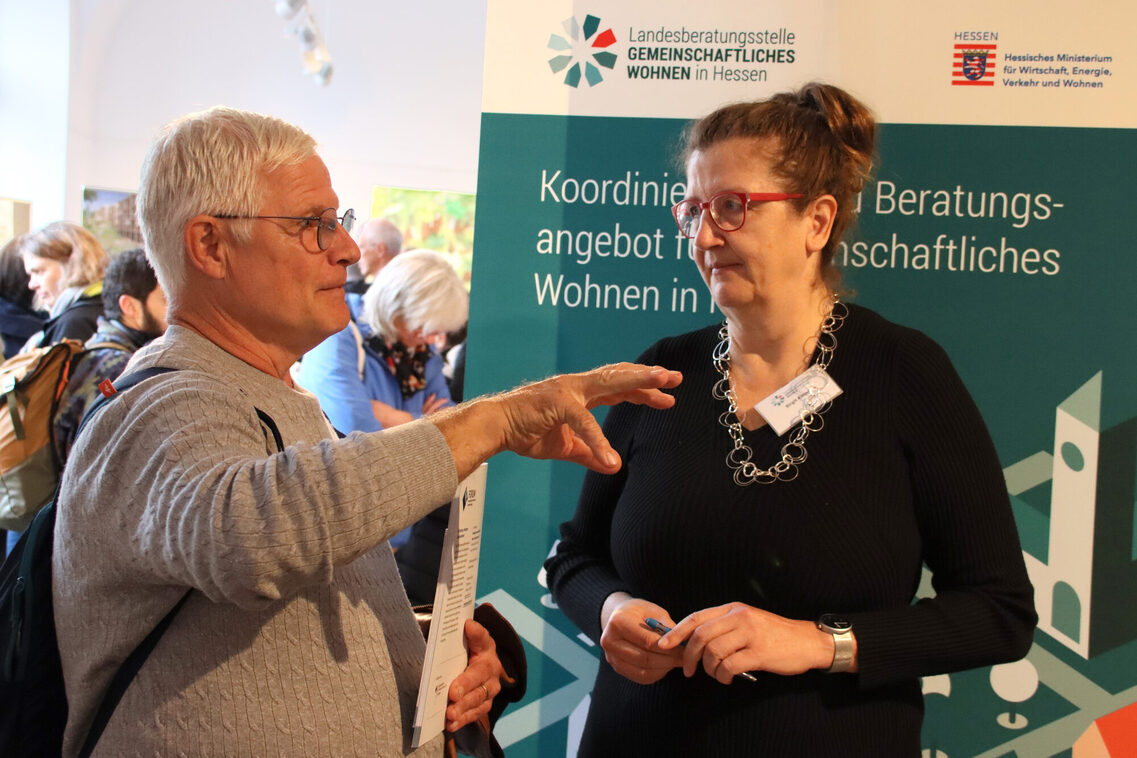 Zwei Personen mittleren Alters unterhalten sich. Eine trägt ein Namensschild. Sie stehen vor einem Banner mit der Aufschrift „Landesberatungsstelle Gemeinschaftliches Wohnen in Hessen“, daneben das zugehörige Logo, außerdem das Logo bzw. Wappen des Landes Hessen mit dem Schriftzug „Hessisches Ministerium für Wirtschaft, Energie, Verkehr und Wohnen“.