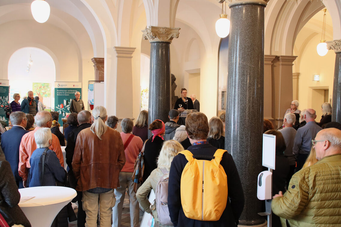 Viele Besucherinnen und Besucher stehen während einer Ansprache in der Säulenhalle des Rathauses, den Rücke zur Kamera. Von der Decke hängen einzelne, ovale Lampen.