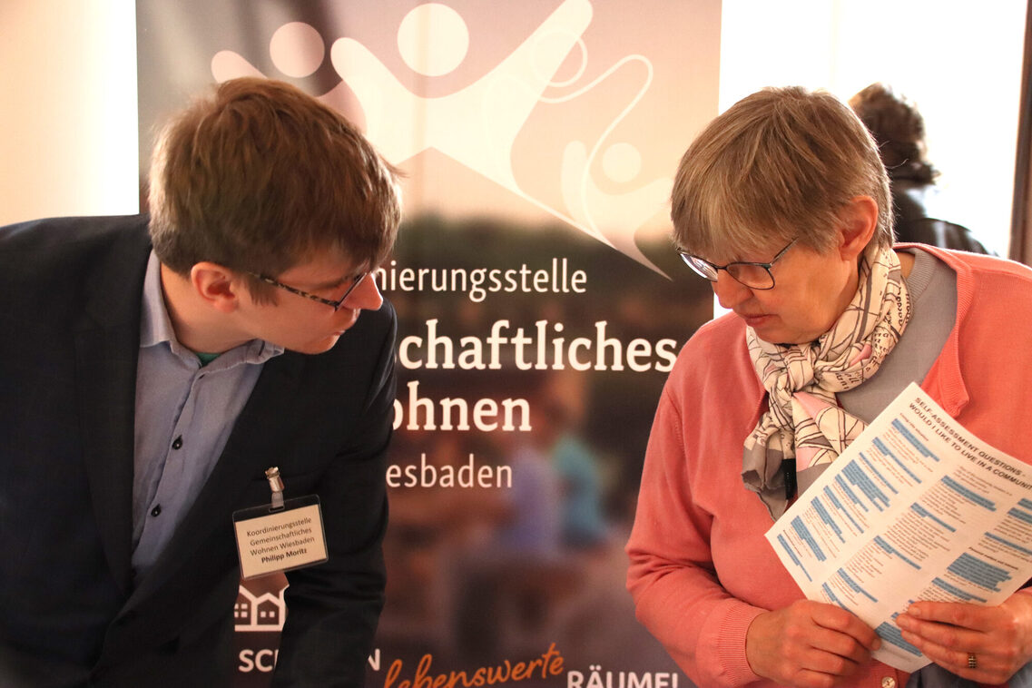 Zwei Personen stehen vor einem Banner mit dem Schriftzug „Koordinierungsstelle Gemeinschaftliches Wohnen in Wiesbaden“. Eine Person trägt ein Namensschild, die andere hält ein bedrucktes Blatt Papier in den Händen.