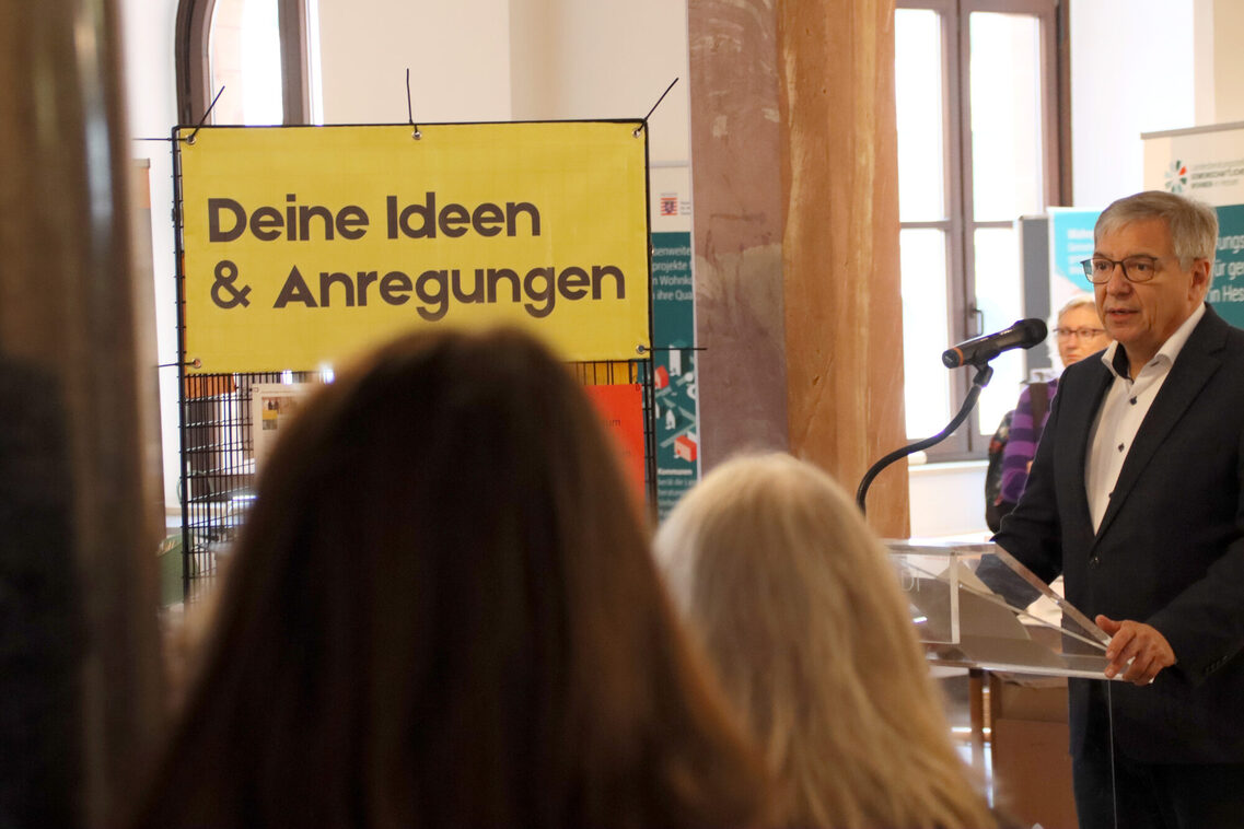 Neben einem gelben Schild imt der Aufschrift „Deine Ideen und Anregungen“ steht Oberbürgermeister Gert-Uwe Mende an einem Rednerpult und spricht in ein Mikrofon. Im Vordergrund sind die Hinterköpfe zweier Personen mit langen offenen Haaren zu sehen.