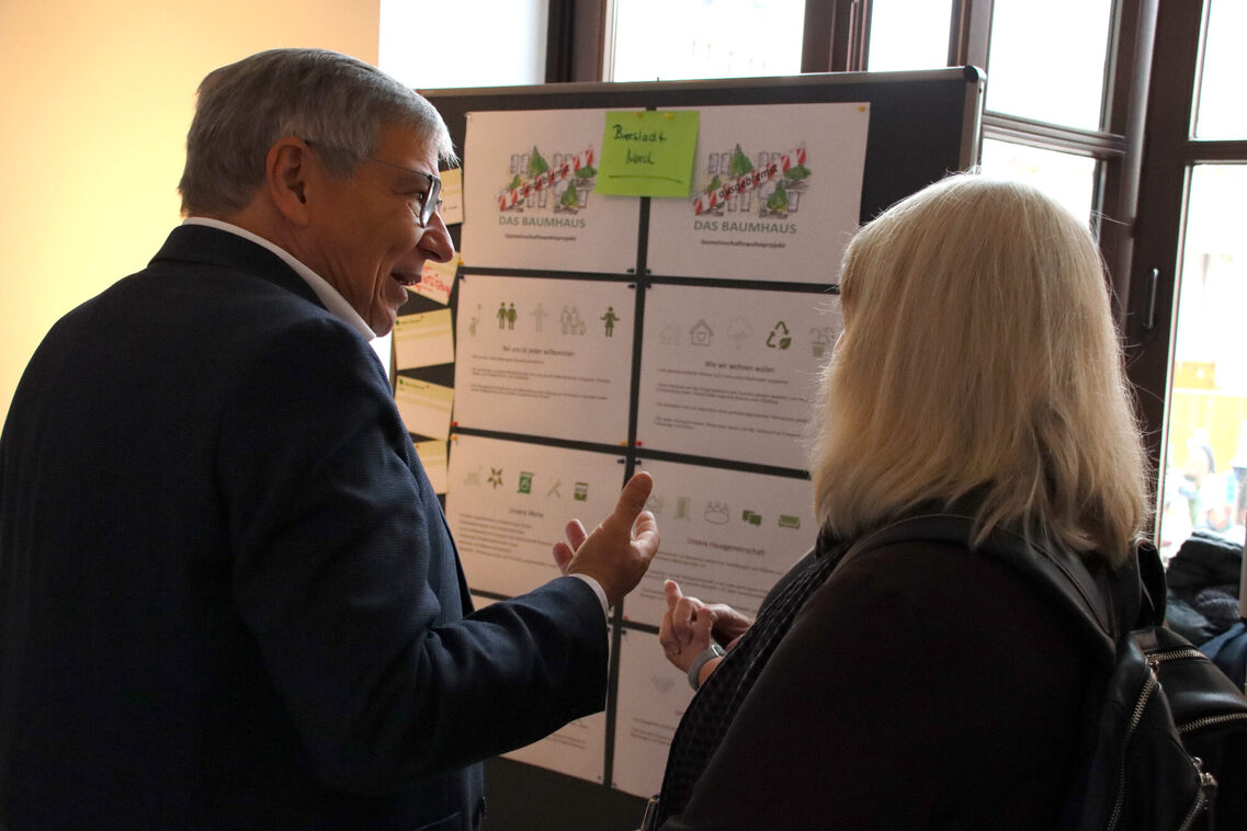 Oberbürgermeister Gert-Uwe Mende steht ins Gespräch mit einer Person vertieft vor einer Pinnwand, an der acht mit Texten und Piktogrammen bedruckte Din A4 Blätter hängen.
