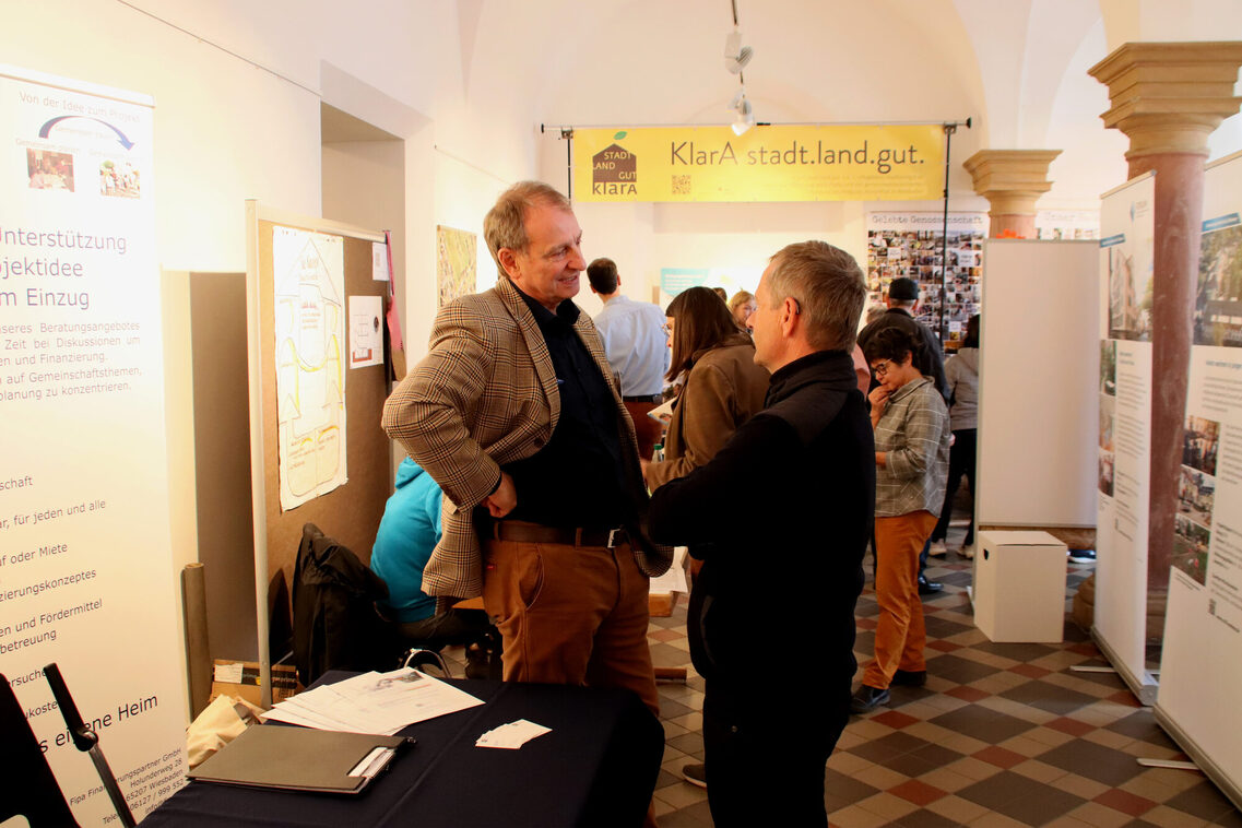 Zwei Männer mittleren Alters stehen ins Gespräch vertieft vor bodentiefen Infobannern in einem Raum mit kariertem Steinboden und Kreuzgewölbe. Im Hintergrund weitere Personen