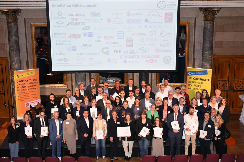 Große Gruppe von Menschen in formeller Kleidung steht in einem Saal vor einer Leinwand mit vielen Logos und hält Urkunden, zwei gelbe Banner mit Text zu ÖKOPROFIT stehen seitlich.