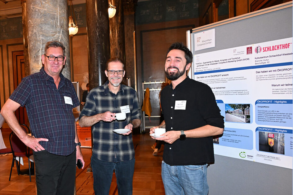 Drei Männer mit Kaffeetassen stehen vor einem Informationsplakat vom Schlachthof.