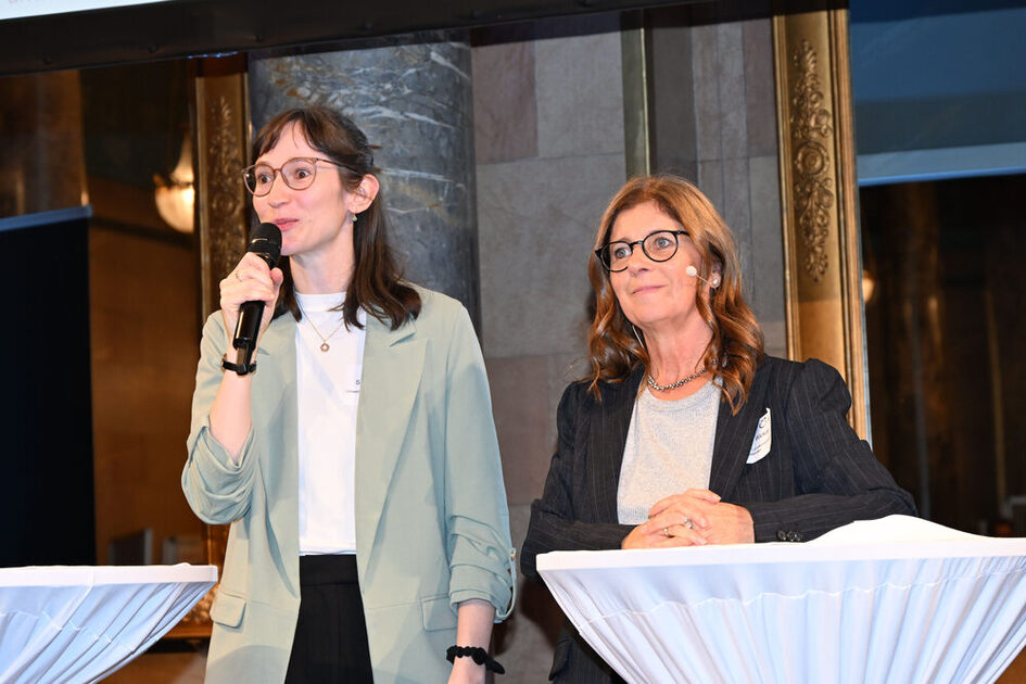 Zwei Frauen stehen an Stehtischen, eine hält ein Mikrofon, beide tragen Blazer und stehen vor einer Säule mit goldverzierten Rahmen.