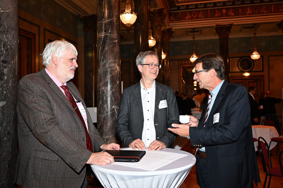 Drei Männer in festlicher Kleidung stehen an einem Stehtisch.