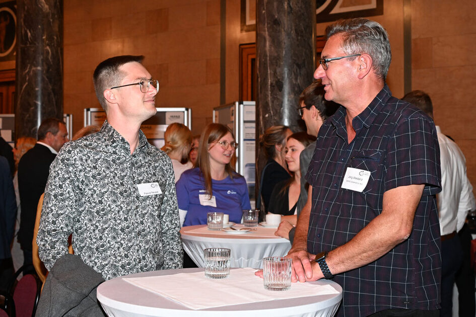 Zwei Männer in gemusterten Hemden stehen an einem Stehtisch in einem holzvertäfelten Raum.