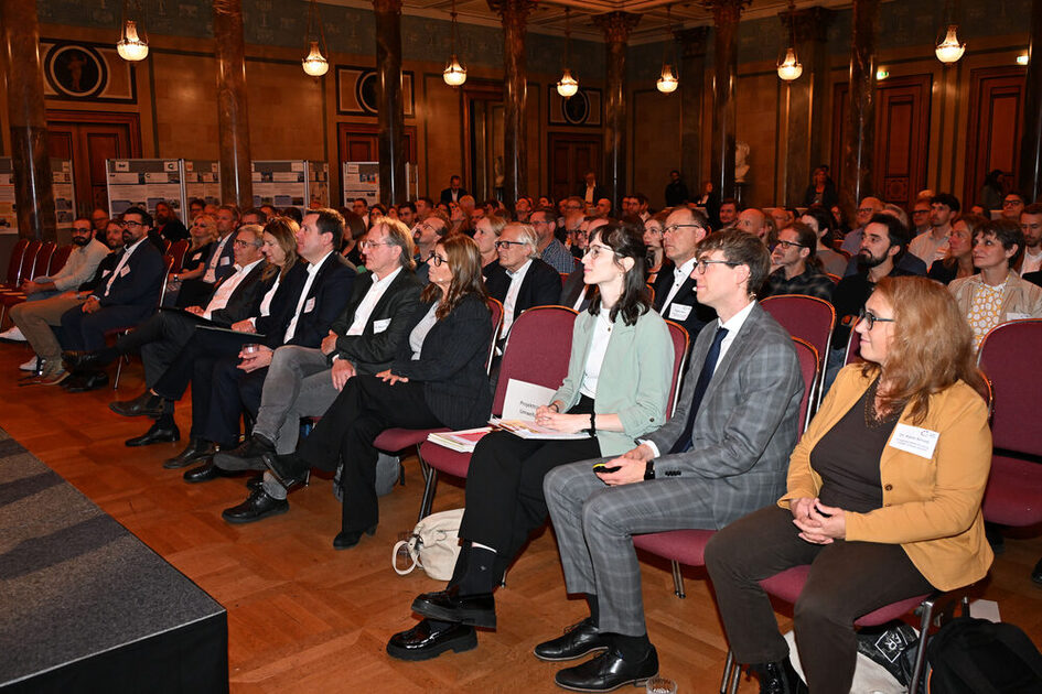 Festlich gekleidete Menschen sitzen auf rot gepolsterten Stühlen, in mehreren hintereinanderliegenden Reihen, in einem holzvertäfelten Saal.