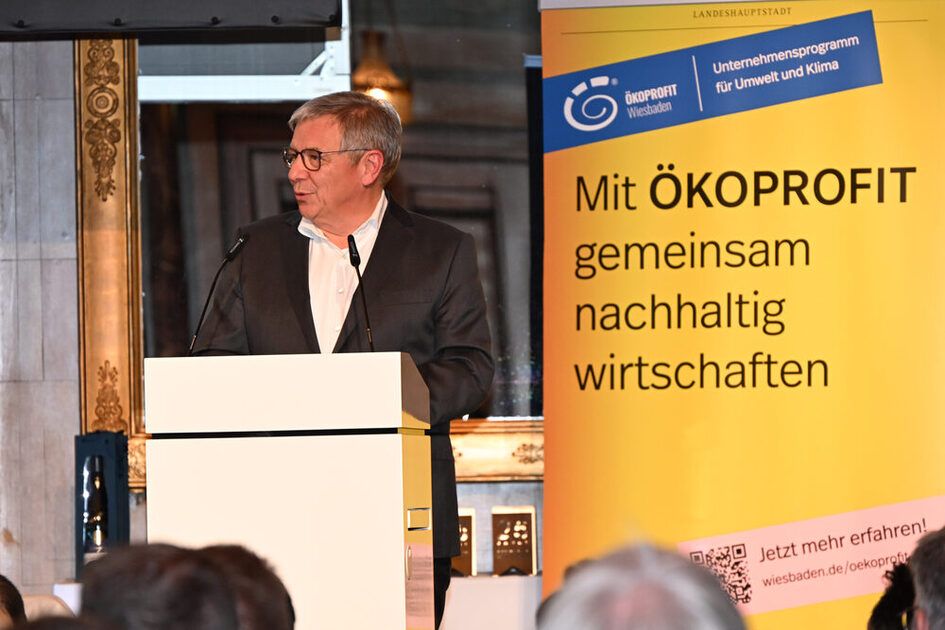 Der Oberbürgermeister von Wiesbaden, Gerd Uwe Mende, steht an einem Rednerpult und hält eine Rede. Neben ihm steht ein Banner mit der Aufschrift Ökoprofit.