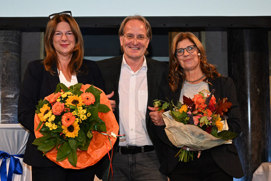 Drei Personen stehen nebeneinander, die beiden äußeren halten große bunte Blumensträuße, alle tragen formelle Kleidung.