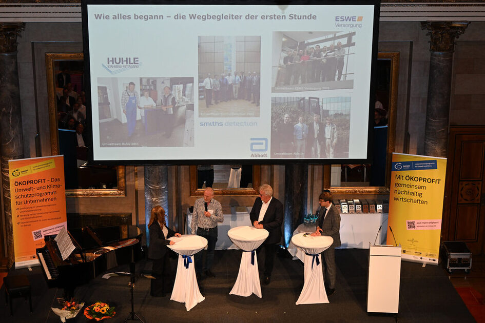 Vier Personen stehen an drei mit weißen Hussen bedeckten Stehtischen auf einer Bühne, im Hintergrund eine Präsentationsfolie mit Fotos und Texten, links und rechts gelbe Banner mit Texten zu ÖKOPROFIT.