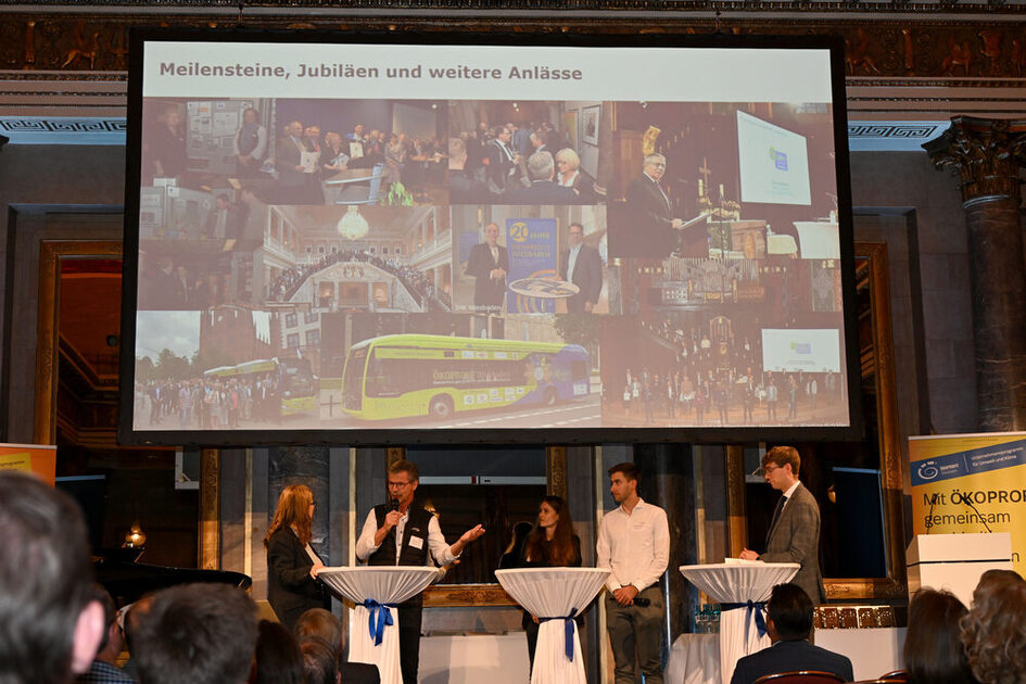 Fünf Personen stehen an drei mit weißen Hussen bedeckten Stehtischen auf einer Bühne, im Hintergrund eine Präsentationsfolie mit Fotos, rechts ein gelbes Banner mit Texten zu ÖKOPROFIT.
