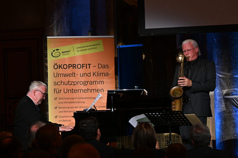 Ein Mann sitzt in dunkler Kleidung an einem schwarzen Flügel, rechts von ihm ein grauhaariger stehender Mann mit Saxophon, hinter beiden steht ein gelbes Banner mit Texten zu Ökoprofit.