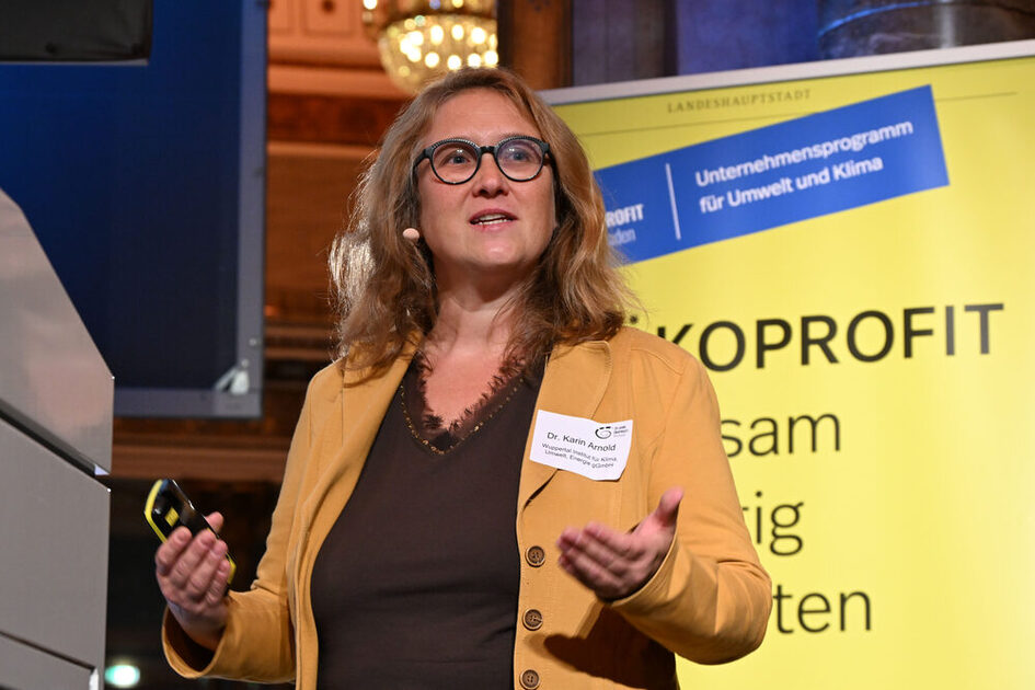 Frau mit braunem Oberteil und gelber Jacke hält Präsentationsfernbedienung, steht vor gelbem Banner mit Text zu Umwelt- und Klimaprogramm.