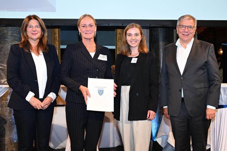 Vier festlich und alle dunkel gekleidete Personen stehen nebeneinander, die mittleren Personen halten eine Urkunde in der Hand.