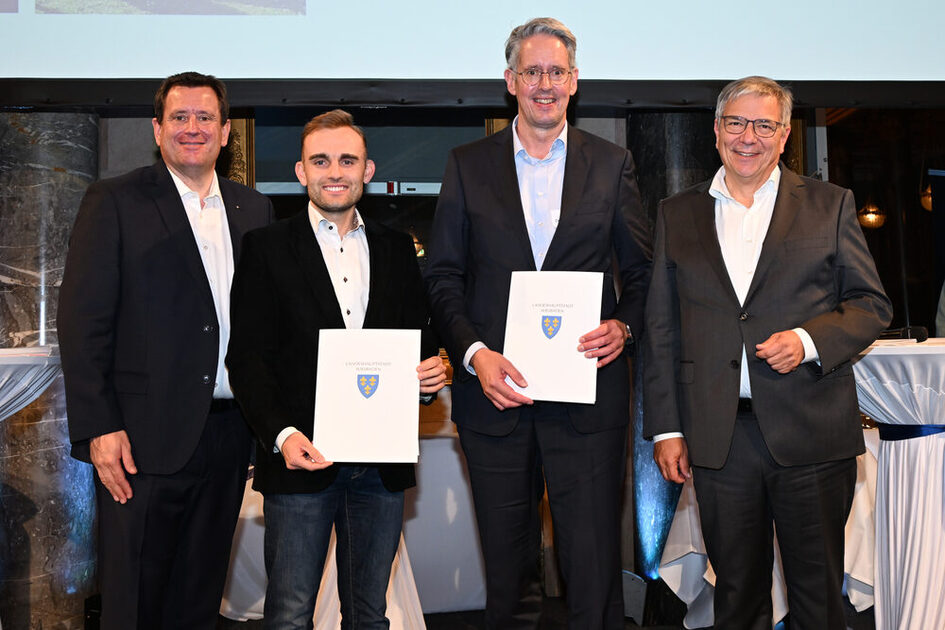 Vier festlich und alle dunkel gekleidete Personen stehen nebeneinander, die mittleren Personen halten Urkunden in der Hand.