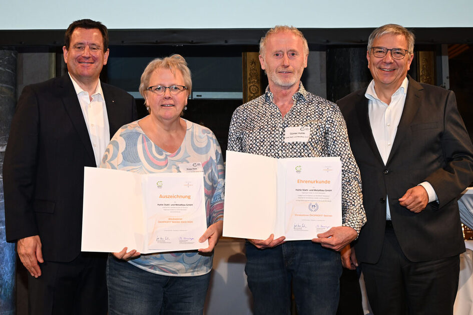 Vier Personen stehen nebeneinander, die beiden mittleren haben Urkunden in der Hand und gemusterte Kleidung. Die Personen links und rechts tragen weiße Hemden und schwarzes Sakko.