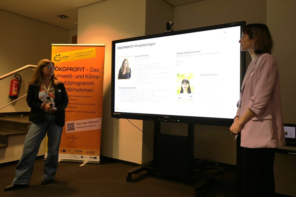 zwei Frauen, zwischen ihnen steht ein helle erleuchteter Bildschirm und ein orangenes Banner mit der Aufschrift ÖKOPROFIT.