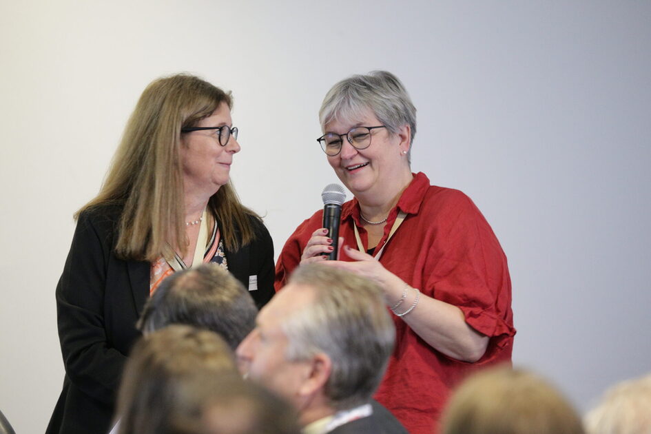 zwei Frauen stehen nebeneinander vor Publikum, eine Frau in roter Bluse spricht in ein Mikrofon während sie von einer Frau in schwarzer Kleidung angeschaut wird.